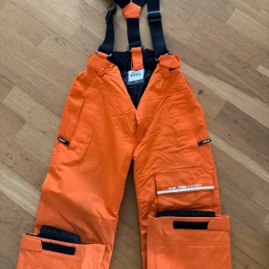 Orange täckbyxor med hängslen - Säljer ett par orange täckbyxor från McKinley med justerbara svarta hängslen. Byxorna har flera praktiska fickor och dragkedjor, perfekt för vinteraktiviteter. De är designade för att hålla dig varm och torr i kalla förhållanden.