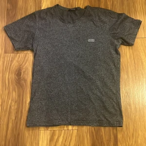 Grå t-shirt från Hugo Boss - Snygg grå t-shirt från Hugo Boss med diskret logga på bröstet. Perfekt för en stilren och avslappnad look. Tillverkad i mjukt material för extra komfort.