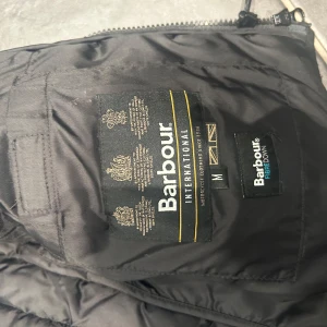 Svart quiltad väst från Barbour - Säljer en svart quiltad väst från Barbour med huva och dragkedja. Västen har flera praktiska fickor med knappar och är perfekt. Den är stilren och funktionell, idealisk för lager-på-lager-stil.