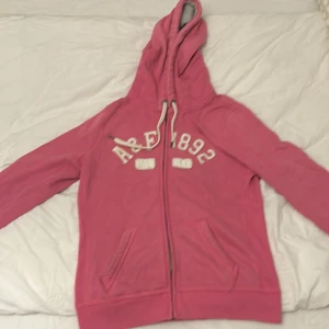 Rosa hoodie från Abercrombie & Fitch - Säljer en snygg rosa hoodie från Abercrombie & Fitch. Den har en dragkedja framtill och en stor, mysig huva med snören. På bröstet står det 'A&F 1892' i vita bokstäver. Perfekt för en avslappnad stil eller kyliga dagar. Passar både till vardags och chillkvällar! jag som har s kan ha den 