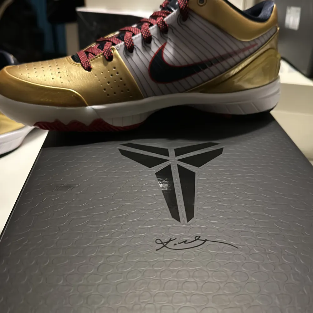 Säljer ett par Nike Kobe IV Protro sneakers i en snygg kombination av guld och mörk obsidian. Skorna har en vit bas med en mörkblå Swoosh och röda detaljer. De är i nyskick och har en cool design med perforerad tå och signaturdetaljer på sidan. Perfekta för både basket och streetwear!. Skor.