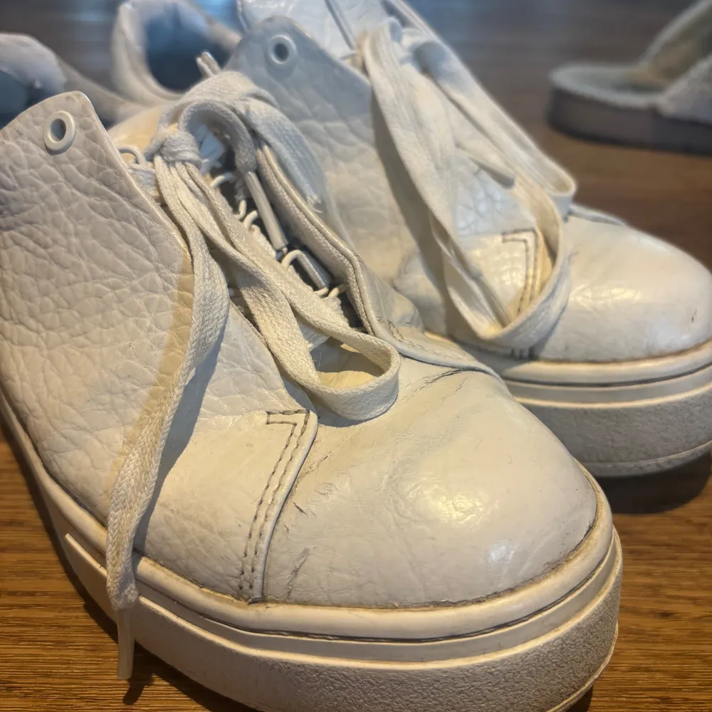 Säljer ett par vita sneakers i skinn. De har en klassisk design med snörning och en tjock sula. Skorna är välanvända med synligt slitage, speciellt vid hälen där materialet har börjat flagna. Perfekta för vardagsbruk om du gillar en avslappnad stil. Dom är väldigt sköna och passar både tjej o kille💕. Skor.