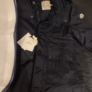 Moncler field jacka - LEZ RAIN JACKET Helt ny! 10/10 skick! Tillverkad av vattenavvisande nylon, Lez-regnjackan för män främjar konceptet med användbarhet med rymliga patchfickor. Perfekt till vår och sommar, hör av dig vid fundering! ALLT OG KOMMER TILL+FYSISKT KVITTO 