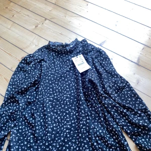 Svart blommig klänning från Zara - Säljer en superfin svart klänning från Zara med små vita blommor. Klänningen har långa ärmar och en hög krage med volangdetaljer. Perfekt för både vardag och fest! Den är i nyskick och har fortfarande prislappen kvar. Passar perfekt till höst och vår.