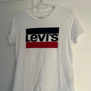Vit t-shirt från Levi's - Säljer en snygg vit t-shirt från Levi's med deras ikoniska logga i svart, blått och rött på bröstet. T-shirten är i bomull och har en normal passform. Perfekt för vardagsbruk eller att matcha med jeans för en avslappnad look. Den är i bra skick och redo för en ny ägare!