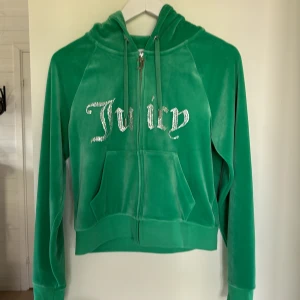 Grön hoodie från Juicy Couture - En grön hoodie från Juicy Couture med dragkedja framtill och en stor ficka. Hoodien har texten 'Juicy' i glittriga stenar på framsidan och en huva med dragsko. Endast använd 1 gång. inomhus.