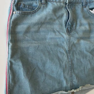 Jeanskjol med detalj på sidan  - Hej, jag säljer en jeanskjol med detalj på sidan med blå, vit och röd färg. Kjolen är sparsamt använd och i fint sick 💕är osäker på vart jag köpt den men tror typ Kappahl. Storleksmässigt skulle jag säga att den är i S/M
