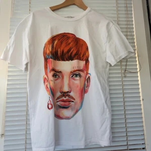 Vit t-shirt med konstnärligt ansikte - En vit t-shirt med ett stort, färgglatt konstnärligt ansikte tryckt på framsidan. Ansiktet har detaljer som piercingar och örhängen, samt texten 'ONE OF MANY' under ögat.