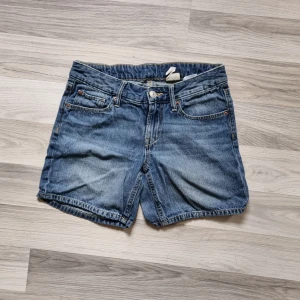 Blå jeansshorts - Ett par blå jeansshorts med fem fickor och knappgylf. Shortsen har en klassisk design med två framfickor, en myntficka och två bakfickor. De är tillverkade i denim och har en något sliten look.