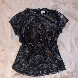 T-shirt  - Svart med skiftning av silver. Gjord i spetstyg så passar bra med svart linne under eller någon färg som lyser igenom.  Guldig Dragkedja i nacken. Aldrig använd bara testad en gång. 