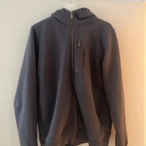 Sail Racing  - En grå huvtröja med dragkedja framtill. Huvan är fodrad med mjukt tyg. Tröjan har två fickor framtill.