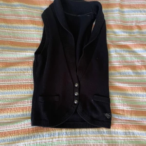 Svart stickad väst - En svart stickad väst med ribbade kanter och tre knappar framtill. Västen har två fickor och en liten dekorativ detalj i form av en siffra 16 i nedre högra hörnet.