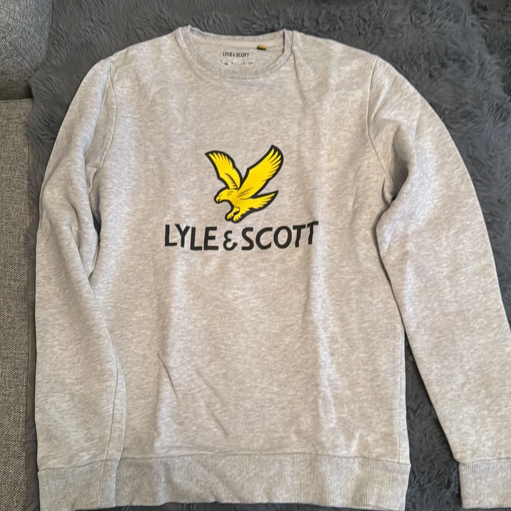 En bekväm grå sweatshirt från Lyle & Scott med ett stort gult fågeltryck och varumärkesnamnet på framsidan. Tröjan har långa ärmar och ribbstickade muddar vid ärmslut och nederkant.. Tröjor & Koftor.