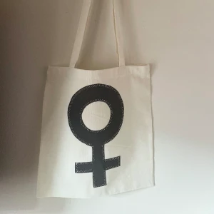 Tygkasse, tygväska, tygpåse  - Handsydd tygkasse med motivet ”feminist”.   Tillverkad i 100% återvunna textilier. Feministtecknet är tillverkat i reflextyg.   Mått: 32x35 cm  Hämtas på Södermalm alternativt skickas med post. 