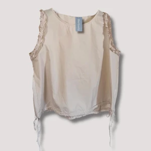 Milli Top  - Ärmlös top med krås i ärmöppningen och knyt i botten på sidorna med dragsko. Söt bas top att antingen bära uppklädd på fest eller lite mer casual till vardags.  One size, passar upp till st. 42  Material: 100% Bomullspoplin   Hör av er vid intresse🌷