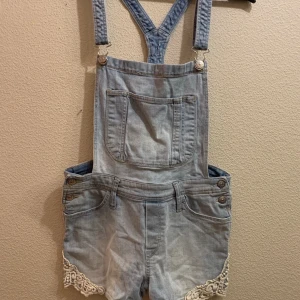 Hängselbyxor / shorts 170, S , XS - Ett par hägselshorts / shorts. Jeans material med fin lite spets broderat dom detalj på benen. Ställbara i hängslen tryckknappar i sidan. St 170 men fungerar enkelt för xs och s.  