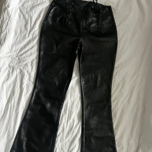 Skinnbyxor i skinnimitation  - Svarta bootcut byxor i skinnimitation med knyte framtill. Oanvända från ASOS. Stl M
