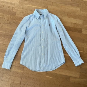 Blå/Vit Ralph Lauren-skjorta - Denna skjorta är perfekt till dig som vill hänga med i trenderna och vill se fräsch ut i sommar. Skjortan är i mycket gott skick då den använts  sparsamt. Kontakta för fler bilder vid intresse:) Priset kan sänkas vid snabb affär👍 Nypris: Ca 1500kr 