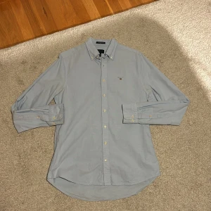 Ljusblå skjorta från Gant - En stilig ljusblå skjorta från Gant i storlek medium. Skjortan har en klassisk krage med knappar och är långärmad. Den är tillverkad i ett bomullsmaterial och har en slim passform. Skjortan är i ett fint skick och har ett luftigt material.