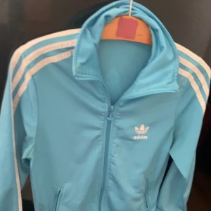 Adidas kofta  - Ljus blå adidas kofta . Storleken ”m” . Finns en liten fläck där bak nära axeln . Använt några gånger 