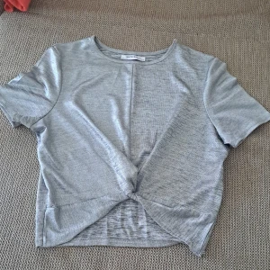 Grå kortärmad topp med knutdetalj - En grå kortärmad topp med rund hals och en knutdetalj framtill. Toppen är tillverkad i ett mjukt och bekvämt material.