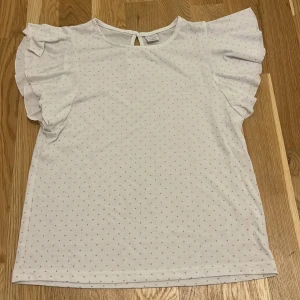 Vit prickig topp från Lindex - En vit topp från Lindex med små bega prickar. Toppen har korta ärmar med volangdetaljer och en knappöppning i nacken.