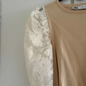 Beige topp från Zara med spetsärmar - En elegant beige topp från Zara med långa spetsärmar i vitt blommönster. Toppen har en rund halsringning och en ribbad nederkant. Änvänd 1 gång. Storlek M men passar även S eller L. 