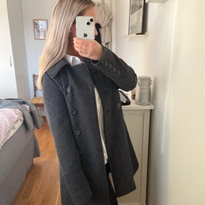 Kappa från Sara Kelly - Ellos  - Mörkgrå kappa från Sara Kelly. Originalpris ca 1000 kr.  Använd vid fåtal tillfällen men ytterst lite nopprig. Går att knäppa hela vägen upp till halsen. Lite liten i storlek. Vid fler frågor, skriv gärna 💕 