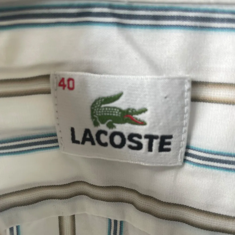 En vit-randig skjorta från Lacoste med logga på bröstfickan | Storlek 40/Medium | Mycket fint skick 👍  Kolla in min profil för fler skjortor, pikétröjor och annat till bra pris! Fler varor kommer inom kort 💸. Skjortor.