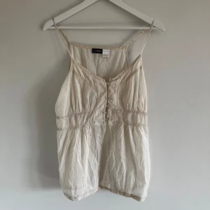 Beige linnetopp från Active Wear - En beige linnetopp från Active Wear med smala axelband och knappar framtill. Toppen har dekorativa sömmar och en lös passform. Är man lite mindre hade den även kunnat sitta som en söt mini klänning💕