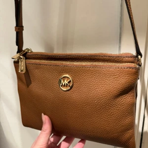 Brun Michael Kors väska crossbody - Brun Michael Kors crosdbody väska med double zip och kortförvaring i mitten. Använd fåtal gånger och inga fläckar inuti. Material är “pebbled leather”. Skriv för fler bilder, har bild på authenticity tag också. Säljer för 750kr eller bud.