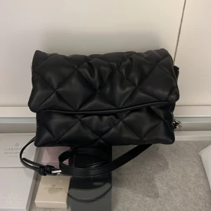 Axelremsväska  - En svart väska med spännbara band. Köpt på Åhléns. 