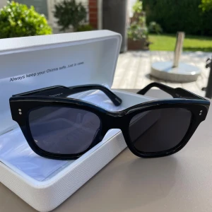 Svarta solglasögon från Chimi - Ett par svarta solglasögon från Chimi med en rektangulär form. Glasögonen har en solid svart färg och kommer i ett vitt fodral. De har små vita detaljer på bågarna nära gångjärnen.