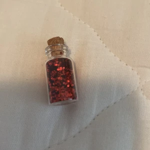 Röd glitterögonskugga i glasflaska - En liten glasflaska med kork innehållande röd glitterögonskugga. Perfekt för att lägga till en glittrande effekt på ögonlocken.