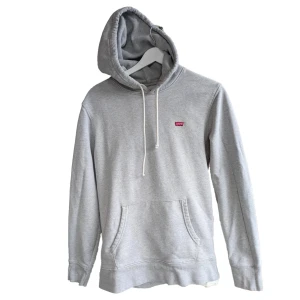 Grå hoodie från Levi's - En bekväm grå hoodie från Levi's med en känguruficka och justerbar huva med vita dragsnören. Tröjan har långa ärmar och ribbstickade muddar vid ärmslut och nederkant.