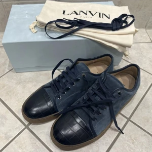 Blå och svarta sneakers från Lanvin - Ett par eleganta sneakers från Lanvin i blå mocka med svarta detaljer. Skorna har en svart tåhätta med ett krokodilmönster och en brun sula. De levereras med en vit förvaringspåse och en blå skokartong.