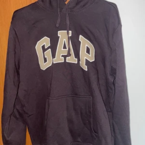 Svart hoodie från GAP - En svart hoodie från GAP med en stor ficka framtill och dragsko vid huvan. GAP-logotypen är tryckt i vitt och grått på bröstet. Tröjan har långa ärmar och en avslappnad passform.