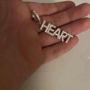 Silverfärgade örhängen med texten 'HEART' - Ett par silverfärgade örhängen med texten 'HEART' dekorerade med små glittrande stenar. Örhängena är långa och hängande, vilket ger en elegant och iögonfallande look.