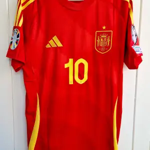 Spaniens matchtröja för sommaren EM  2024 med Pedri på ryggen. Tröjan är en reprint i nyskick med emblem och patchar av högsta kvalité. Storlek: Large. Tröjan är en player version vilket är den tröja som spelarna själva använder. Tajt i storleken.