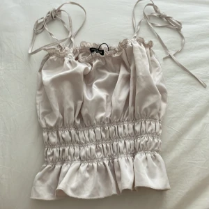 Beige satinlinne med volangdetaljer - Ett beige linne i satinliknande material med justerbara axelband och volangdetaljer vid nederkanten. Linnet har en smockad midja som ger en fin passform.