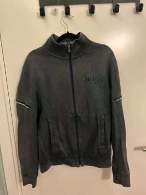 Mörkgrå ziptröja från BOSS - Snygg mörkgrå ziptröja från BOSS med hög krage och diskret logga på bröstet. Tröjan har ribbade muddar, fickor framtill och detaljer på ärmarna. Perfekt för en avslappnad men stilren look. Koftan är ganska missfärgad och har ett par hål/skador lite här o där. Se bilder