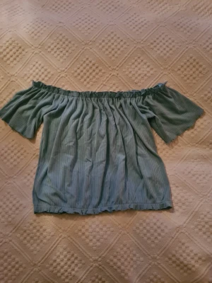 Blå/grön offshoulder topp från Bik Bok - Superfin blå/grön offshoulder topp från Bik Bok med ribbad struktur. Toppen har korta ärmar och en smockad resår upptill som håller den på plats. Perfekt att styla med jeans eller kjol för en trendig look.