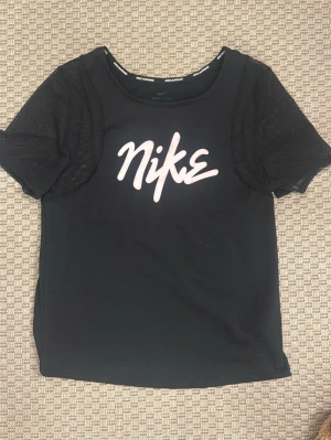 Svart Nike t-shirt med meshärmar - Snygg svart t-shirt från Nike med vit logga i graffiti-stil på bröstet. Tröjan har meshärmar som ger en sportig vibe och är tillverkad i ett lätt, funktionsmaterial. Perfekt för dig som gillar streetwear och sportiga outfits.