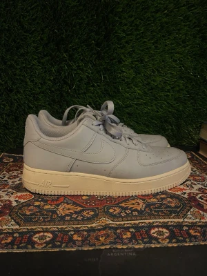 Nike Air Force 1 låga sneakers grå - Snygga Nike Air Force 1 sneakers i ljusgrått läder med klassisk vit sula. Ikonisk swoosh på sidan, perforerad tåbox och vadderad plös för extra komfort. Perfekta för dig som gillar stilrena och tidlösa sneakers.