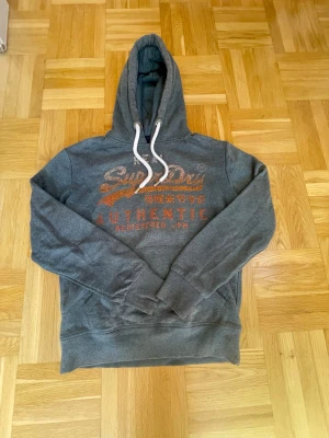 Grå Superdry hoodie  - Snygg grå hoodie från Superdry med orange och vitt tryck på bröstet där det står 'Superdry Authentic'. Hoodien har vit dragsko i huvan och en klassisk känguruficka framtill. Perfekt för en avslappnad och cool stil.