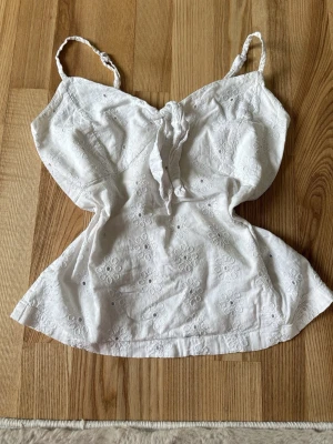 Vit broderad topp med knyt - Söt vit topp med broderade blommor och knyt framtill. Axelbandslös modell med smala axelband och lätt utsvängd passform. Perfekt för varma dagar och sommarens alla äventyr.