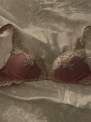 Rosa spets-bh med beige detaljer - Snygg bh i dammig rosa med vacker beige spets över kuporna och axelbanden. Kuporna är lätt vadderade och har en rundad form. Spetsen ger en romantisk och feminin känsla. Perfekt för dig som gillar fina detaljer och mjuka färger.