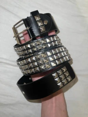 Black Studded Belt - Snyggt svart bälte med flera rader fyrkantiga silverfärgade nitar. Perfekt accessoar för att ge outfiten en edgy vibe. Bältet har klassiskt spänne i metall och är gjort i ett skinnliknande material.