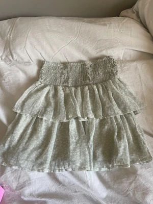 Ljusgrön volangkjol med smock - Supersöt ljusgrön kortkjol med volanger och smock i midjan. Kjolen har ett diskret prickigt mönster och är gjord i ett lätt, luftigt material som passar perfekt till varmare dagar. Den har en flowy passform och ger en riktigt fräsch vibe.
