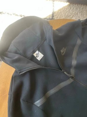 Svart Nike Tech Zip - Tjena👋 Säljer en svart hoodie från Nike med hel dragkedja framtill och huva👌 Hoodien har diskreta svarta detaljer över bröstet och en liten Nike-logga på vänster sida👍 Tillverkad i mjukt material och har långa ärmar samt fickor på sidorna🙏 meddela gärna vid frågor📩